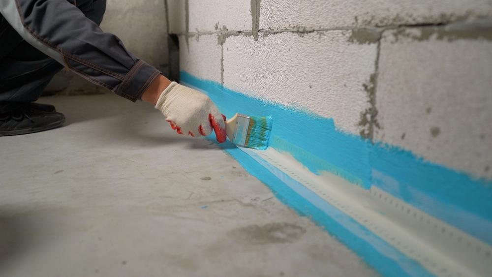 Construction worker waterproofing concrete wall using blue paint and brush in unfinished basement