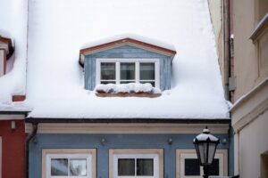 Snow gently blankets the rooftop of a colorful house, revealing a quaint attic window. The winter atmosphere creates a peaceful and picturesque scene.